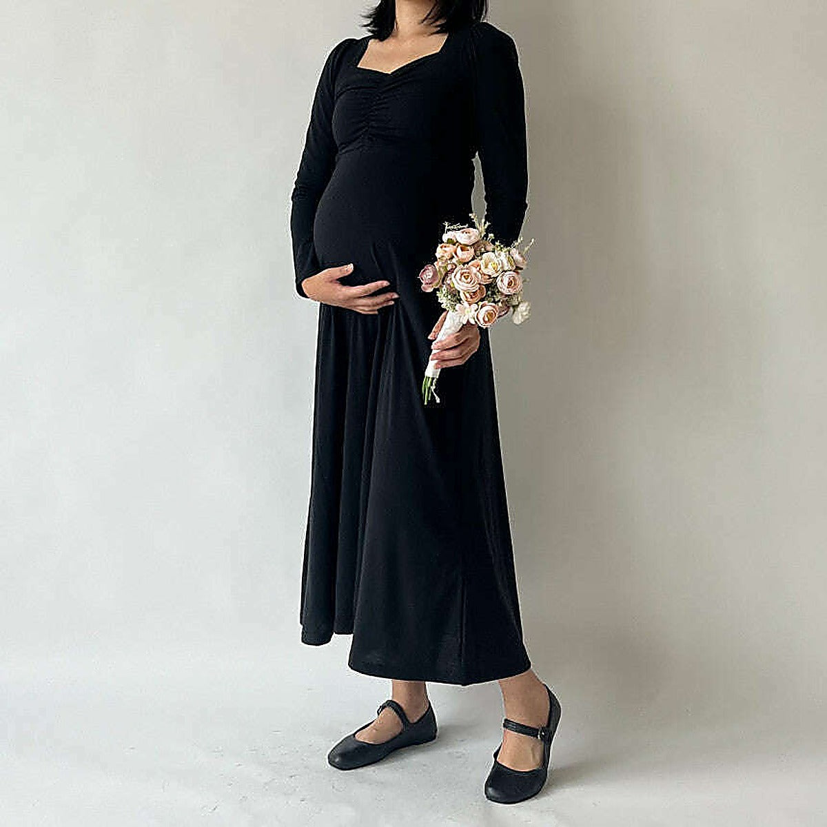 模特手持花束，黑色長裙外搭黑色外套，淺色攝影棚背景｜孕婦心領抽褶舒適連身長裙（第 20 張）